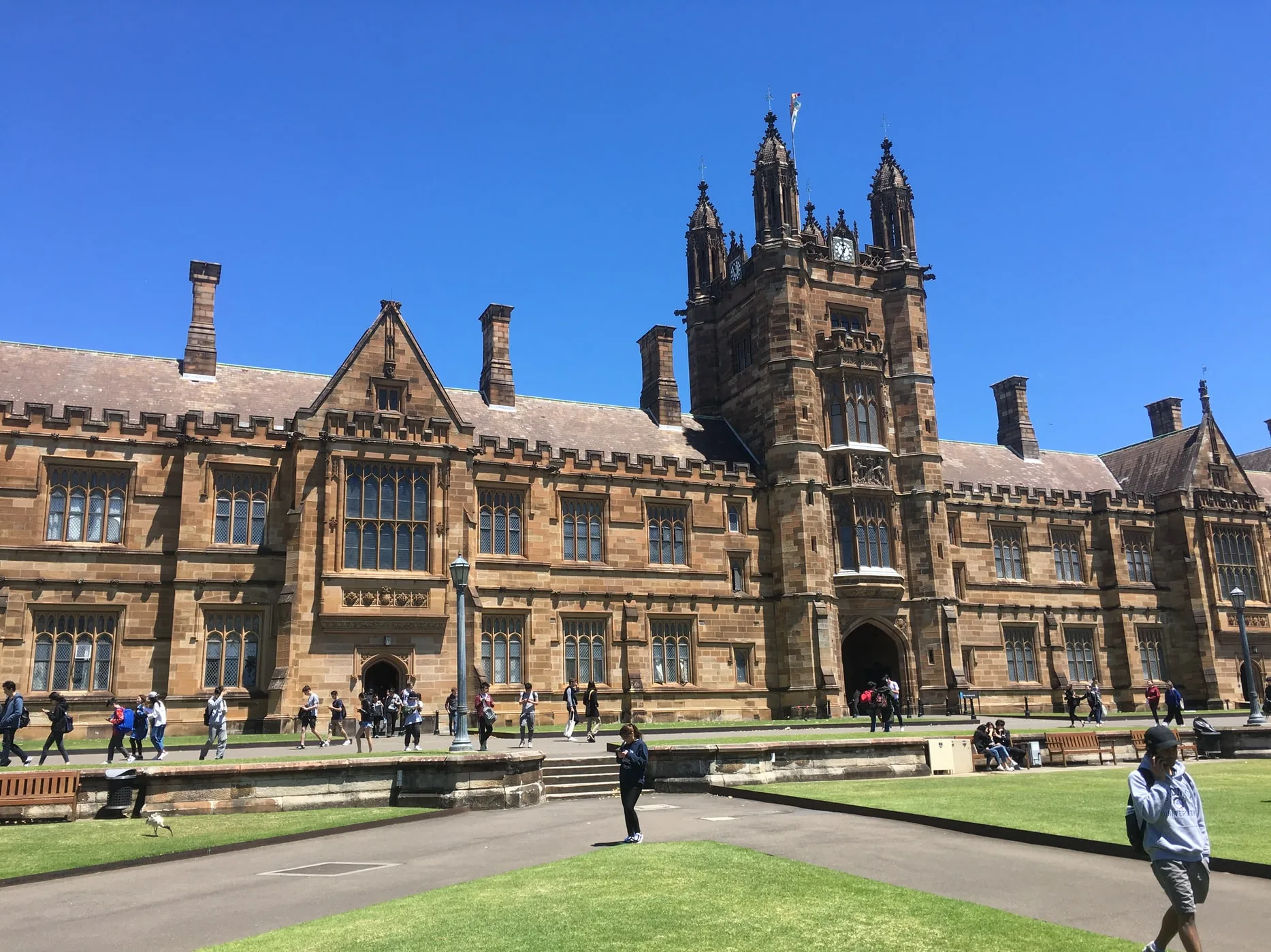 Students walking across a lawn in front of a sandstone university quadrangle building on a sunny day.