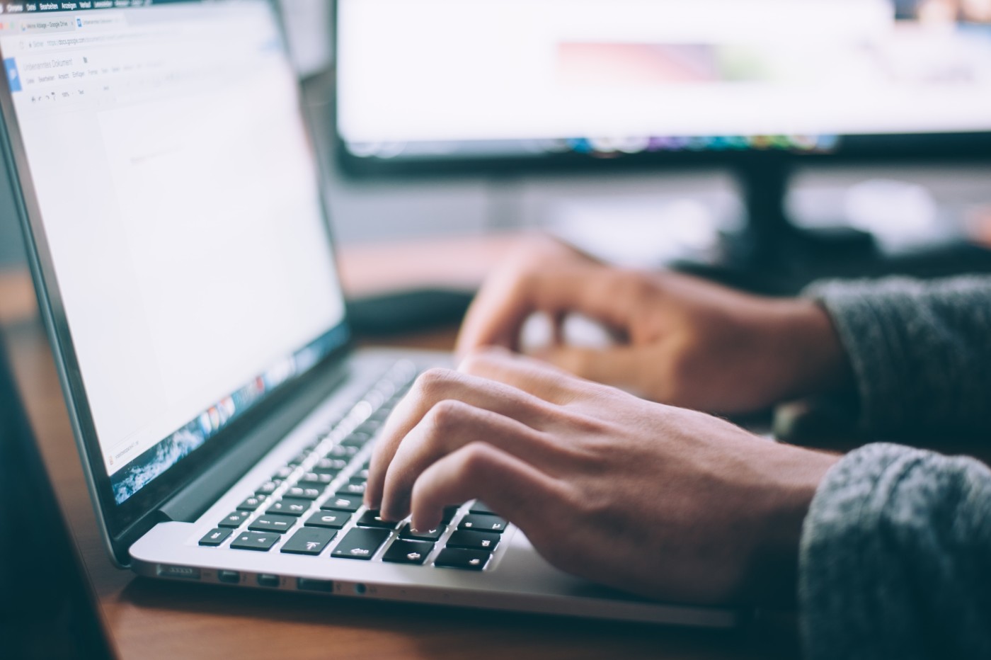 Close-up of hands typing on a laptop keyboard, with a desktop monitor behind it.